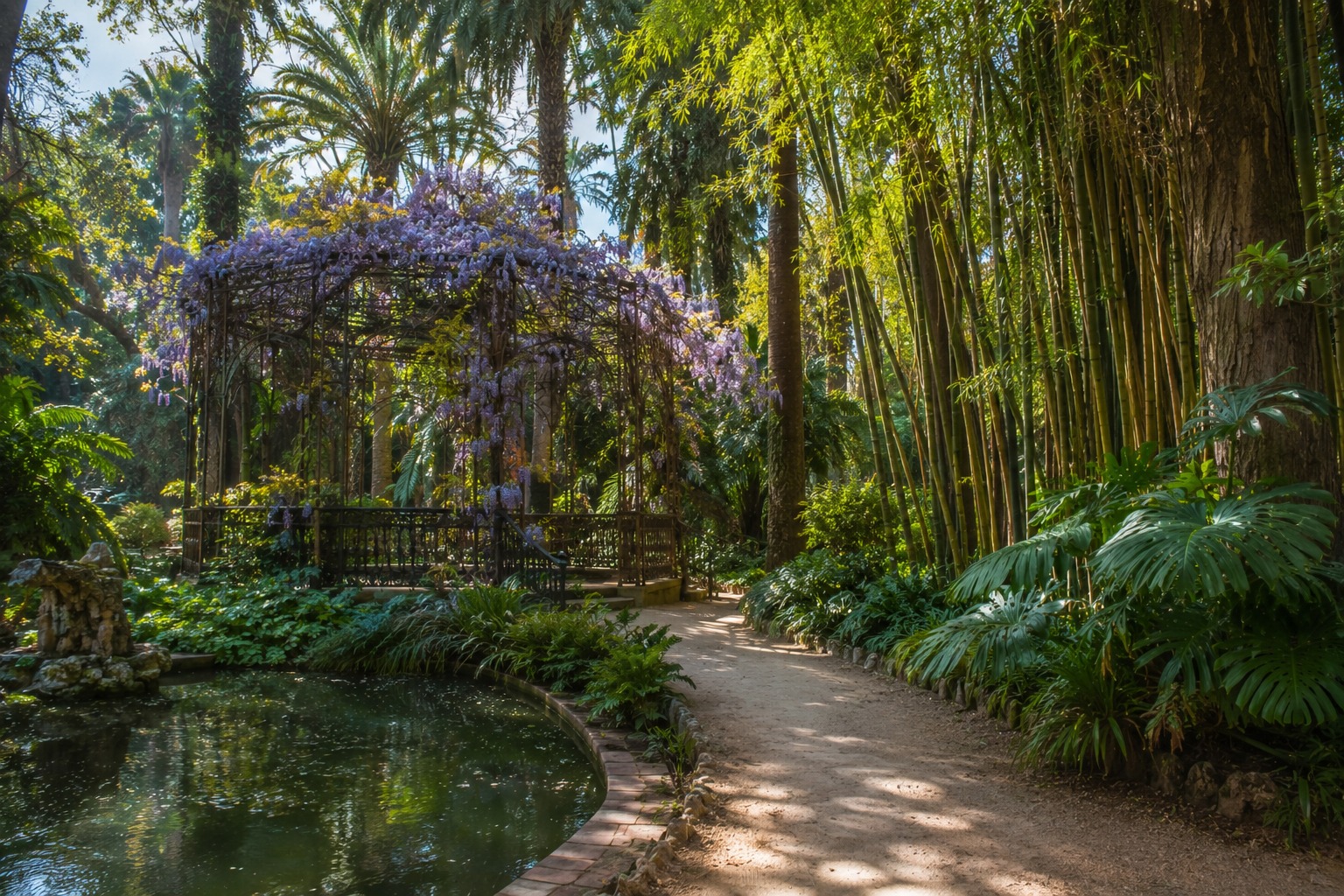 Järngazebo med blommande blåregn vid en damm i La Concepción botaniska trädgård, Málaga — tropiska palmer och bambu omger stigen